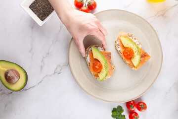 Healthy breakfast toasts with avocado, cherry tomato and smoked salmon on seeded bread. A woman&rsquo;s hand grabs one slice on a neutral plate. Marble background and fresh toppings