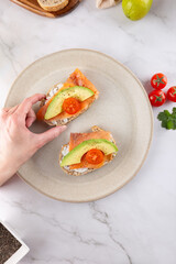 Close-up of a female hand grabbing one toast from a plate. Healthy breakfast with avocado, cherry tomatoes and smoked salmon on whole grain bread. Marble background and fresh ingredients. 