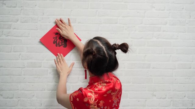 Happy young Asian woman wearing traditional red cheongsam dress decorating home with lucky couplets banner for Chinese New Year festival celebration