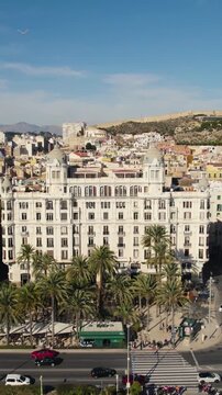 Vertical aerial drone footage capturing Alicante&rsquo;s Casa Carbonell and the palm tree-lined promenade along the Spanish coastline.