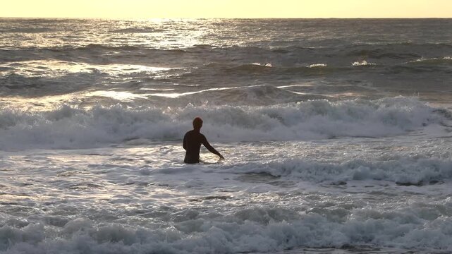 Surfer in warm wetsuit enter water with board at sunset. Winter sports in Italy. Health, sea, and nature.