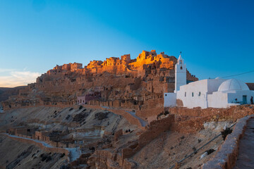 Chenini ksar at golden hour in southern Tunisia, ancient Berber village with white mosque and stone houses on desert hill, dramatic light, heritage, travel destination