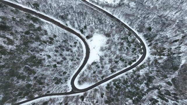 Aerial view of a sinuous road winding through a snow-laden forest, the dark asphalt contrasting starkly with the white trees, Orschwiller, Grand Est, France.