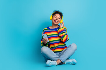 Young woman with rainbow striped sweater listening to music on her phone and smiling against a blue...