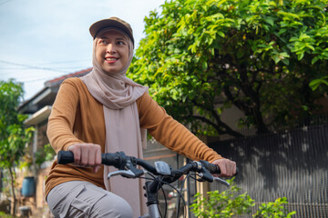 Obraz premium Asian Muslim woman wearing a hijab rides a bicycle along a tree-lined street, enjoying a sunny day.