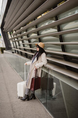 Woman with suitcase and tote leaning on modern building