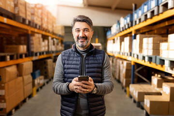 Middle-aged warehouse manager using smartphone in storage. Professional businessman in distribution center. E-commerce, supply chain and technology concept. Logistics worker.