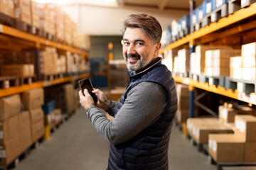 Warehouse worker holding smartphone blank screen mockup. Logistics manager using app for inventory tracking. Digital transformation in industry and supply chain background.