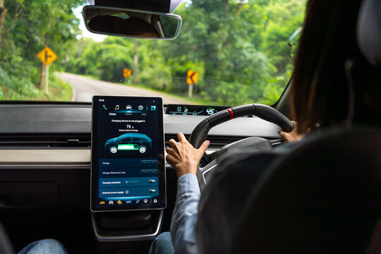 Asian woman driver electric car along a scenic winding road, surrounded by lush greenery. The digital touchscreen displays the electric car battery level and charging options. Future energy concept. - Powered by Adobe