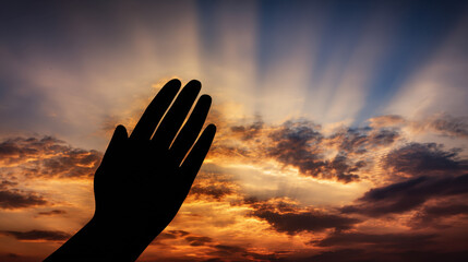 Silhouette of a hand raised in prayer against a sunset sky.