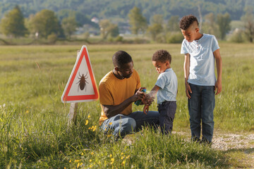 Man taking precautions against insects during a nature walk, spraying his sons near a tick...