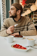 A man sits at a cafe table with cappuccino and desserts, holding a fork and looking away from the window in a cozy modern interior.