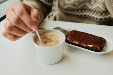 A hand stirs foamy cappuccino in a white cup beside a tiramisu dessert served on a plate in a minimalist cafe setting.