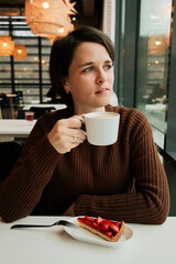 A woman holds a cup of cappuccino near a window with a strawberry tart on the table in a modern cafe interior.