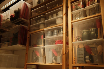 Wooden shelving with transparent plastic containers holding kitchen items, textiles, and decor in a storage room with warm indoor lighting.