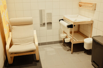 Clean baby care room with a changing table, chair, paper dispenser, and tiled walls, designed for comfort and hygiene in public spaces.