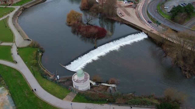 Aerial view of the weir creating a cascade of white water contrasting with the dark river, green park pathways, and a distant green bus, Derby, England, United Kingdom.