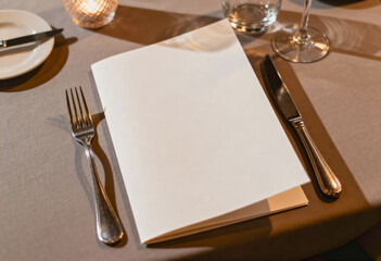 A blank folded menu set on a restaurant table with a textured tablecloth, accompanied by a fork, knife, empty wine glass, water glass, and a lit candle casting warm shadows