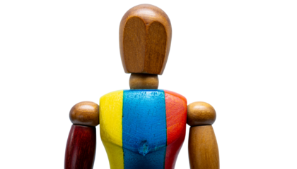 A wooden mannequin figure with a painted torso of red blue and yellow stands against a stark white background in a studio setting