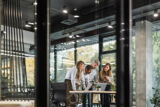 Group of business people standing together and discussing their work and projects, having a team meeting in an office, view from glass wall