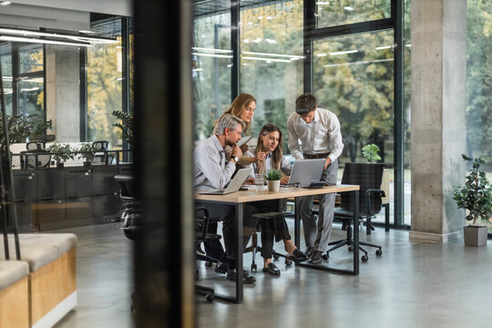 A view through a glass partition shows a modern, busy office interior. Business people are working at desk