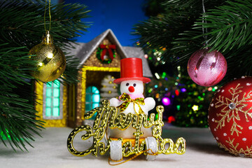 Between two balls on the Christmas tree branches is a snowman on a sled with letters against the background of a house