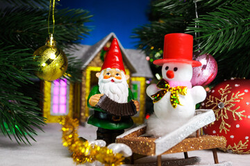 Between two balls on the Christmas tree branches there is a gnome with a snowman on a sled against the backdrop of a house