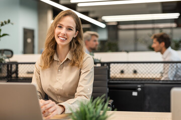 Attractive young businesswoman sitting on the table, looking at camera with colleagues on the background