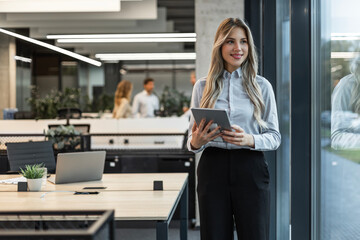 Young elegant business woman standing with digital tablet near office window