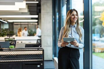 Young elegant business woman standing with digital tablet near office window