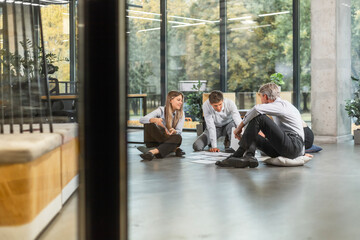 Business people looking at project plan laid out on floor, discussing new project plan in modern office, view from glass wall