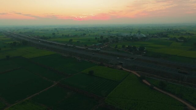 Aerial view of a highway cutting through green fields at sunrise, the landscape a mix of nature and infrastructure, Alwar, Rajasthan, India.