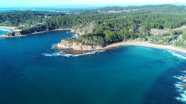 Drone aerial landscape showing clear ocean reef waves crashing along coastline headland covered by National Park trees at Mckenzies Beach rural town suburbs Malua Bay Australia travel tourism nature