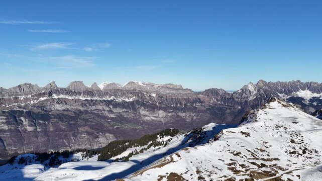 A majestic winter panorama of the Swiss Alps. Crisp blue skies over the snowy slopes of Flumserberg, with the iconic jagged peaks of Churfirsten towering over the Flums valley and Walensee.