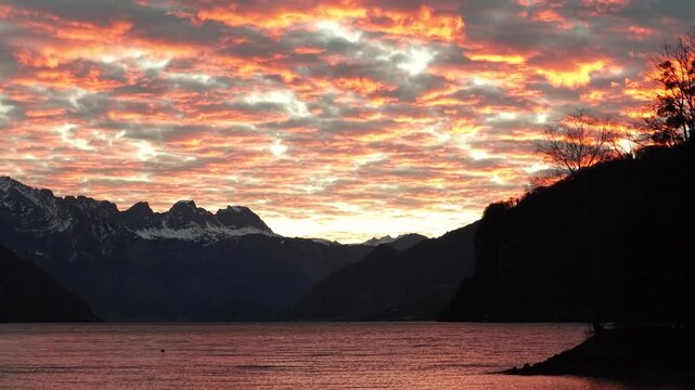 sunrise illuminates clouds above Walensee, with silhouettes of the Alps and shoreline. Switzerland