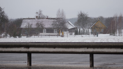 Snow gently falls on wooden buildings in a rural setting. Trees stand bare in the freezing weather. The scene is viewed through a car window on a frigid day
