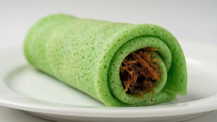 Close-up of a vibrant green rolled pancake, a traditional Indonesian dessert, filled with sweet grated coconut and palm sugar, presented on a white plate.