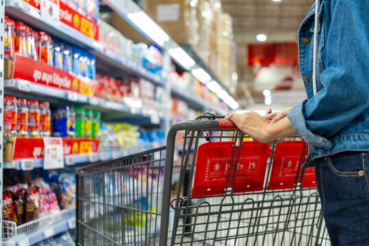 Image of asian woman shopper pushing a red shopping cart in a warehouse store aisle, surrounded by shelves full of bottled water and other items ready for customers to add to their carts.