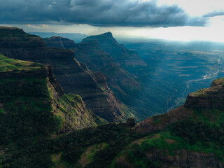 Aerial view of dramatic sunlight piercing through the clouds, illuminating the rugged cliffs and verdant valleys of the Western Ghats, Bhandardara, Maharashtra, India.