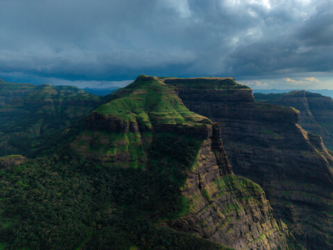 Aerial view of dramatic, rugged mountains cloaked in vibrant green vegetation under a moody, cloud-streaked sky, Bhandardara, Maharashtra, India.