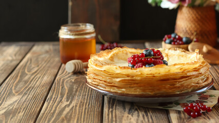 Delicious stack of thin pancakes served with fresh berries and honey on wooden table with flowers in background