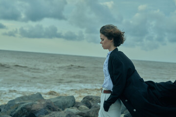 Windy coastal scene of a woman on rocks by the sea, coat billowing behind, calm expression, looking...