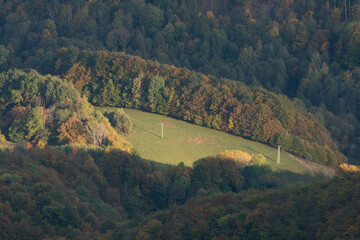 Aerial view of a sun-kissed clearing surrounded by a tapestry of autumn colors and dense forest, with distant poles standing tall, Muranska Planina, Banska Bystrica Region, Slovakia.