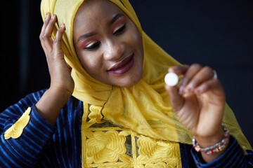 Muslim woman thoughtfully examining a single pill, wearing a yellow headscarf, with a look of concern on black background