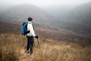Obraz premium Happy hiker in warm clothing standing in nature with backpack and hiking poles, enjoying a peaceful moment in winter wilderness.