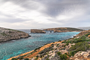Malta Blue Lagoon landscape. Stunning view of the Blue Lagoon in Comino, Malta, showing the famous turquoise crystal clear waters and the rocky islet of Cominotto under a cloudy sky.