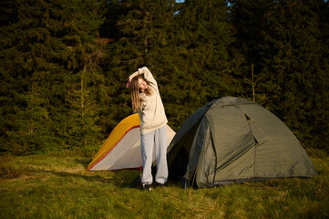 Woman hiker stretching at tent's entrance, warming up in the morning. Young female traveler at the tent looking out at a scenic view. Solo travel, adventure, and enjoying the outdoors.