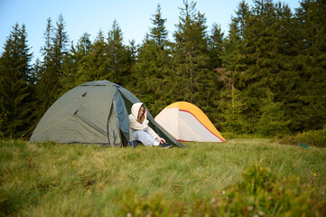 Woman hiker sitting at tent's entrance relaxing in the morning. Young female traveler at the tent looking out at a scenic view. Solo travel, adventure, and enjoying the outdoors.