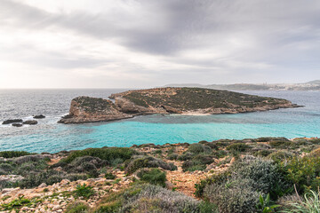 Malta Blue Lagoon landscape. Stunning view of the Blue Lagoon in Comino, Malta, showing the famous turquoise crystal clear waters and the rocky islet of Cominotto under a cloudy sky.