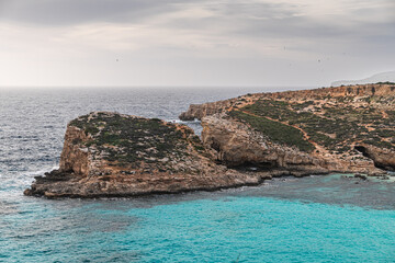 Malta Blue Lagoon landscape. Stunning view of the Blue Lagoon in Comino, Malta, showing the famous turquoise crystal clear waters and the rocky islet of Cominotto under a cloudy sky.